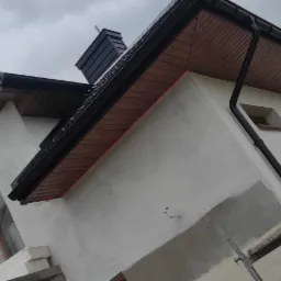 Dom w trakcie prac wykończeniowych elewacji i dachu: widoczne rusztowania, ocieplony budynek z elementami drewnianej podbitki dachowej, czarne rynny i komin, pochmurne niebo.