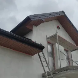 Dom w trakcie prac wykończeniowych elewacji, widoczne rusztowanie, zabezpieczone okna folią, czarne rynny i podbitka dachowa z drewnianą boazerią, pochmurne niebo w tle.