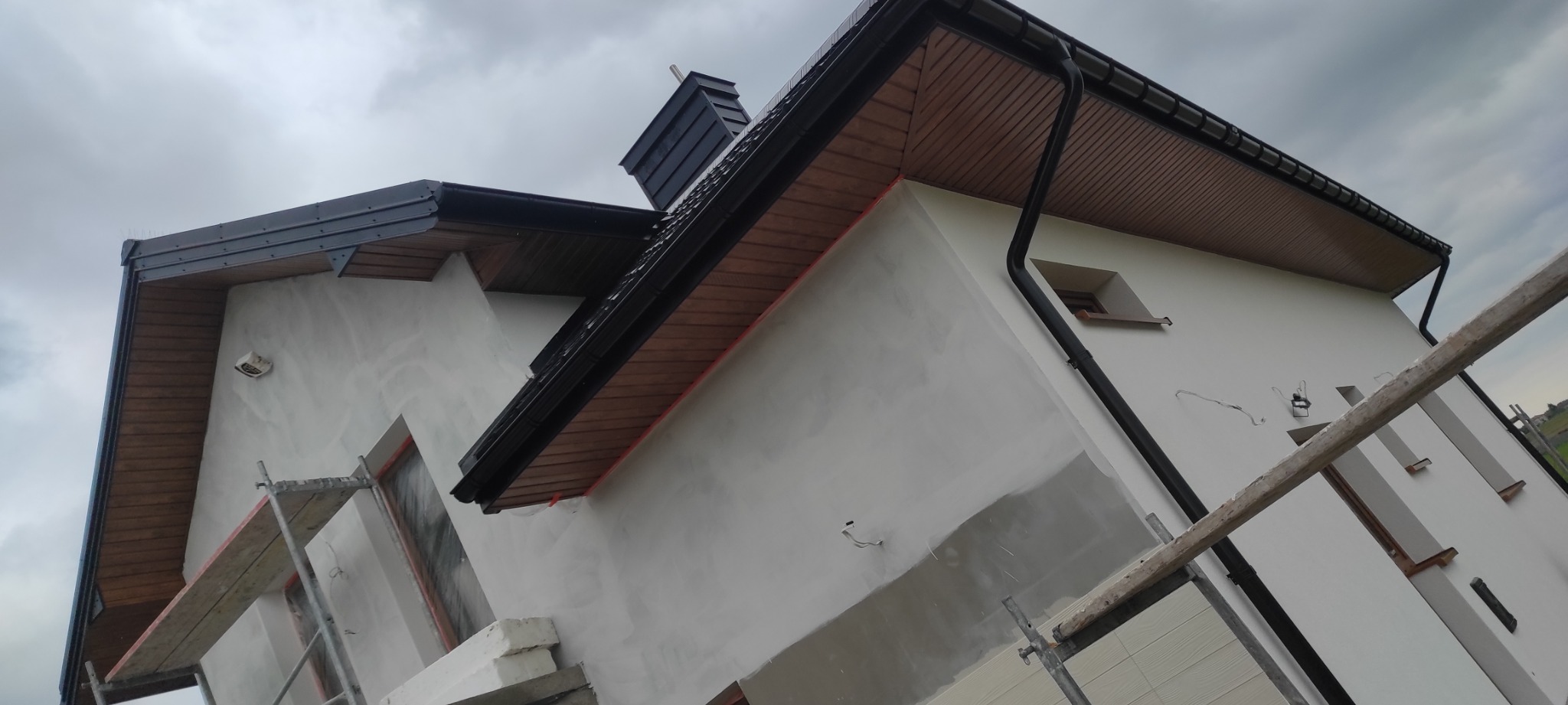 Dom w trakcie prac wykończeniowych elewacji i dachu: widoczne rusztowania, ocieplony budynek z elementami drewnianej podbitki dachowej, czarne rynny i komin, pochmurne niebo.