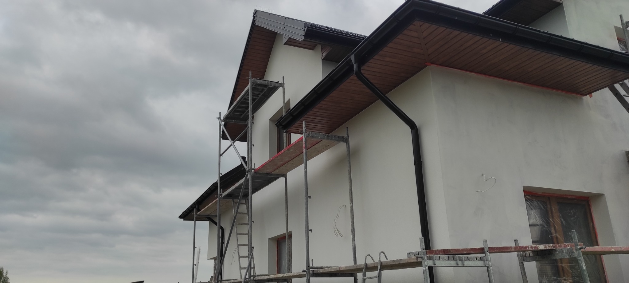 Dom jednorodzinny w trakcie prac wykończeniowych elewacji, widoczne rusztowania, zabezpieczone okna folią, zamontowane rynny w kolorze grafitowym, podbitka drewniana w kolorze brązowym.