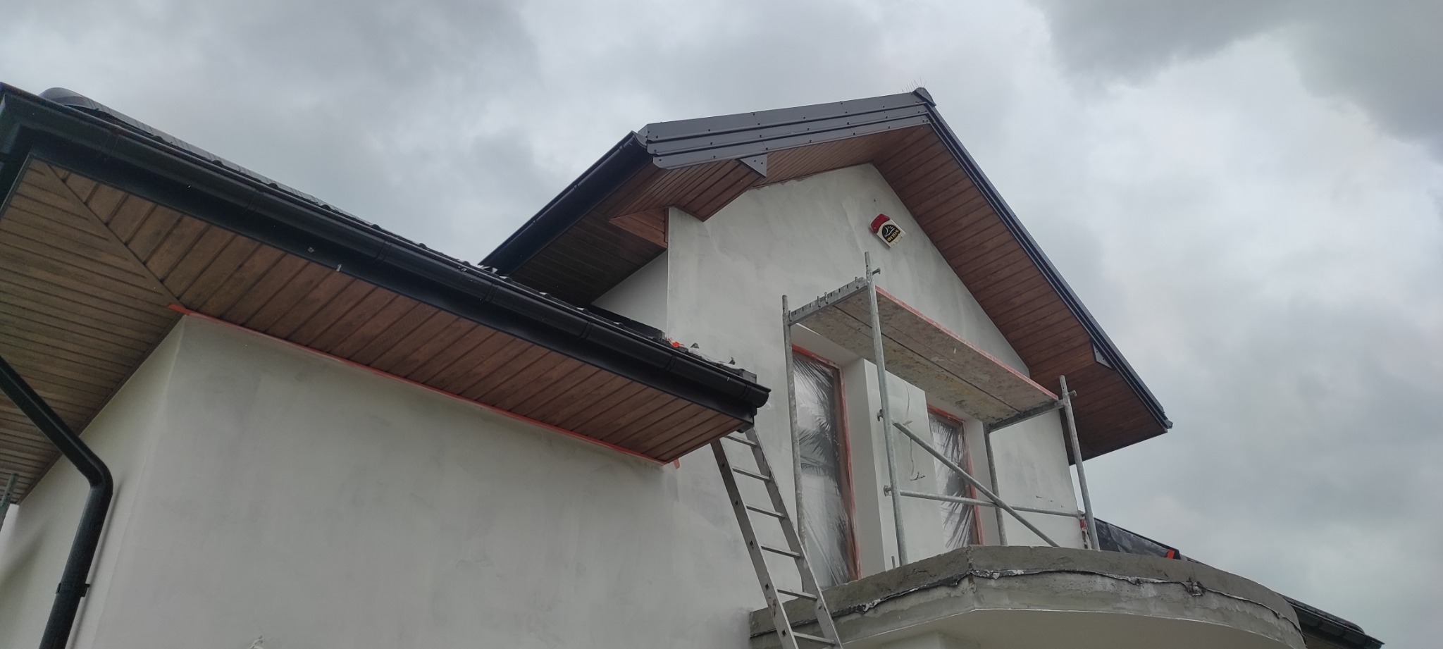 Dom w trakcie prac wykończeniowych elewacji, widoczne rusztowanie, zabezpieczone okna folią, czarne rynny i podbitka dachowa z drewnianą boazerią, pochmurne niebo w tle.