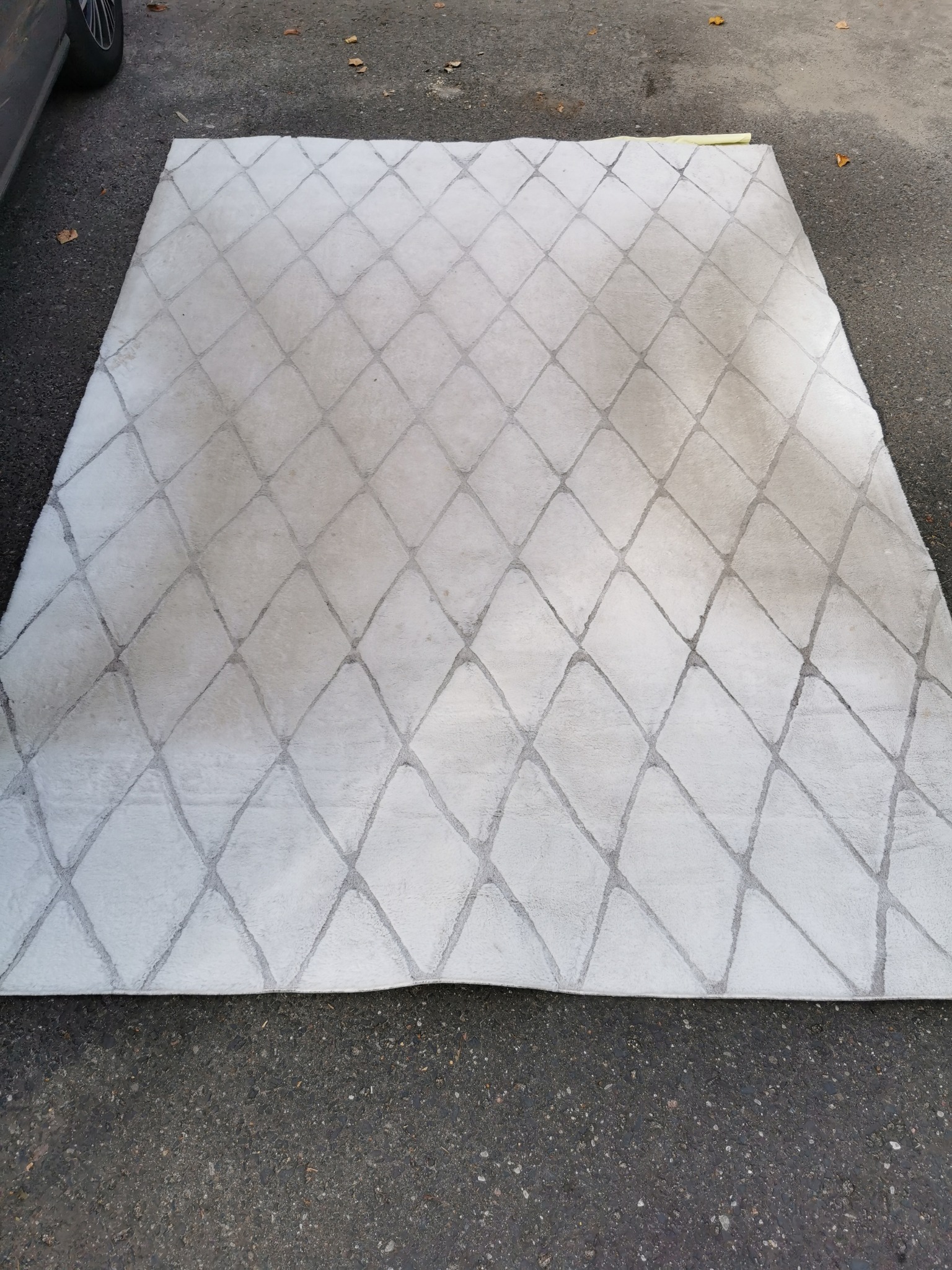 Jasnoszary dywan z geometrycznym wzorem rombów leżący na zewnątrz, widoczne ślady użytkowania i zabrudzenia.