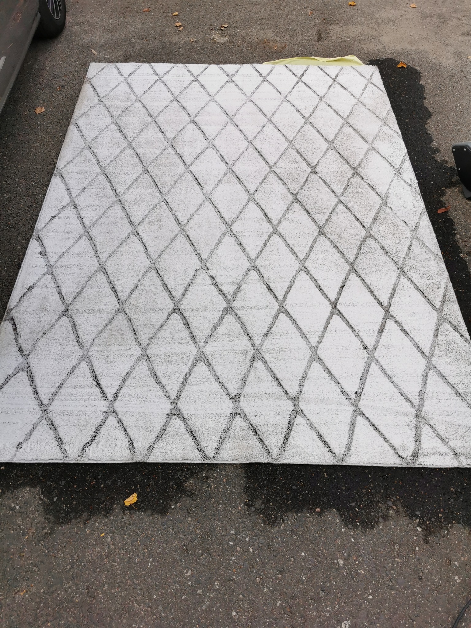 Duży, jasnoszary dywan z geometrycznym wzorem rombów, leżący na mokrym asfalcie, prawdopodobnie po czyszczeniu.