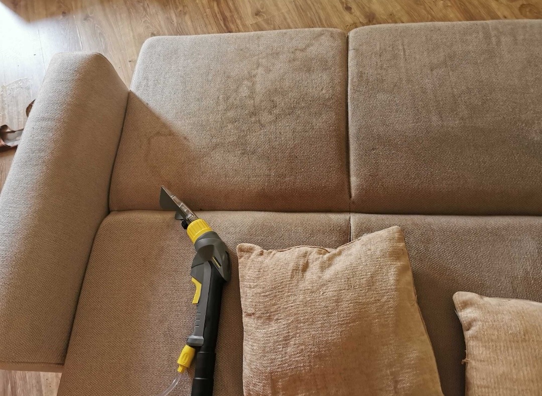 Beżowa sofa z widocznym lekkim zabrudzeniem, czyszczona za pomocą szaro-żółtego odkurzacza piorącego z przezroczystą końcówką, obok leżą dwie poduszki w podobnym odcieniu.