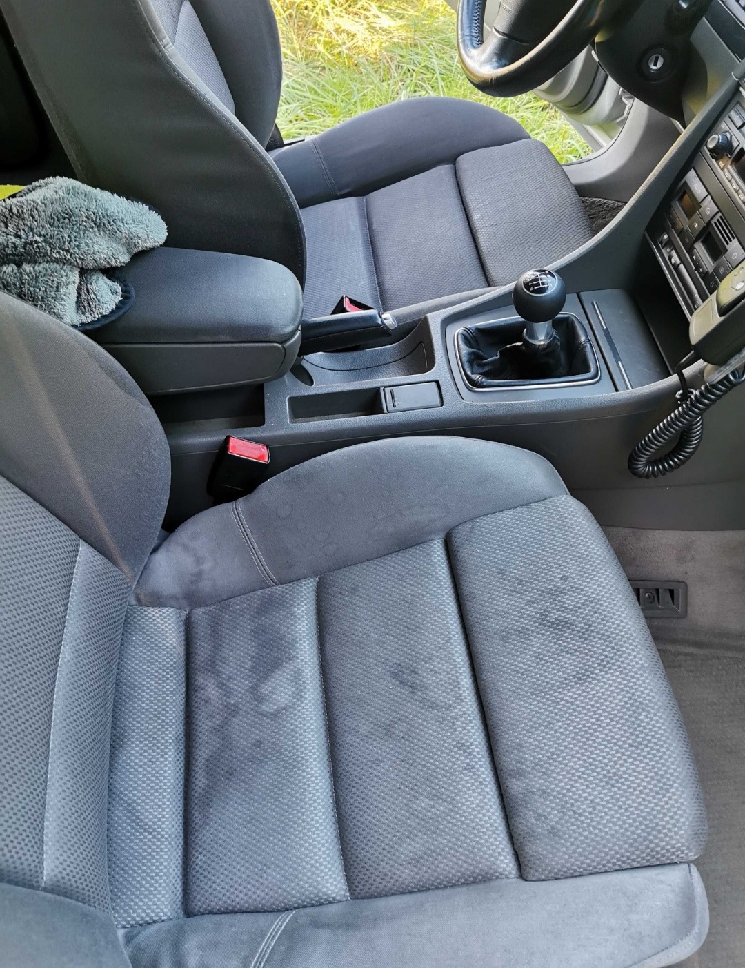 Wnętrze samochodu z szarą tapicerką, widoczne plamy na fotelu kierowcy, dźwignia zmiany biegów i ściereczka z mikrofibry na podłokietniku.