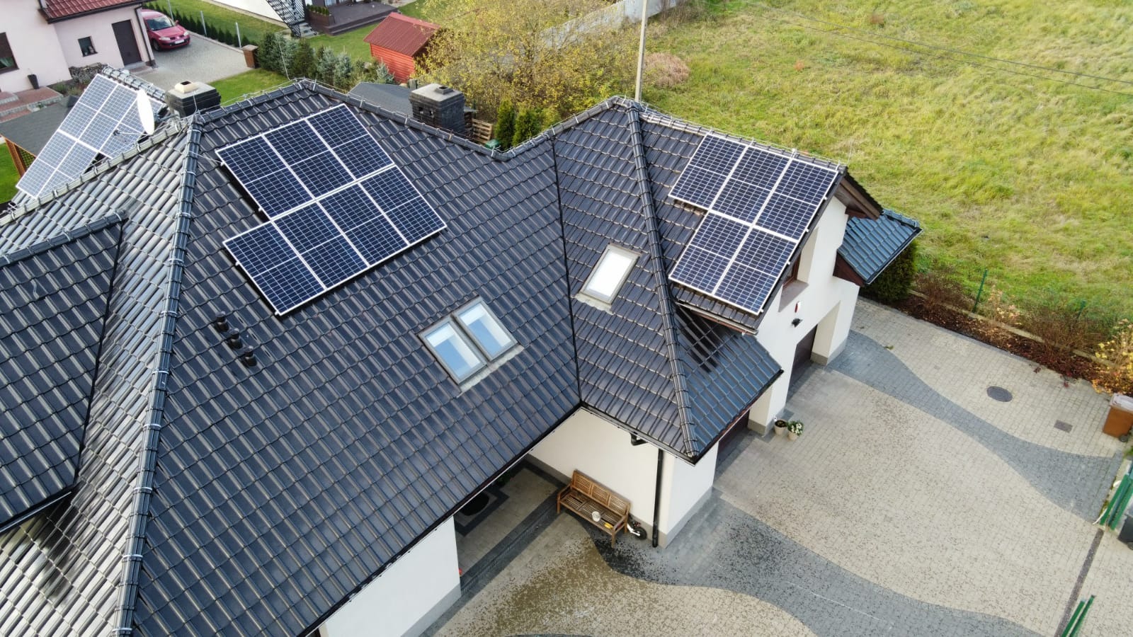 Widok z góry na dom z ciemną dachówką i panelami słonecznymi na różnych połaciach dachu, widoczne okna dachowe i podjazd z kostki brukowej.