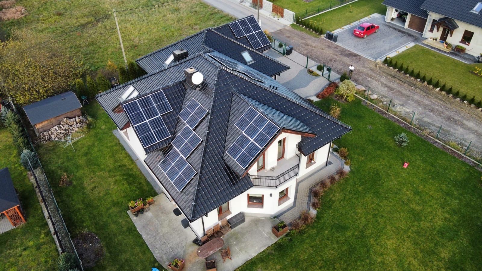Widok z drona na dom jednorodzinny z ciemnym dachem pokrytym panelami słonecznymi, zadbany trawnik, podjazd z kostki brukowej i czerwony samochód w tle.