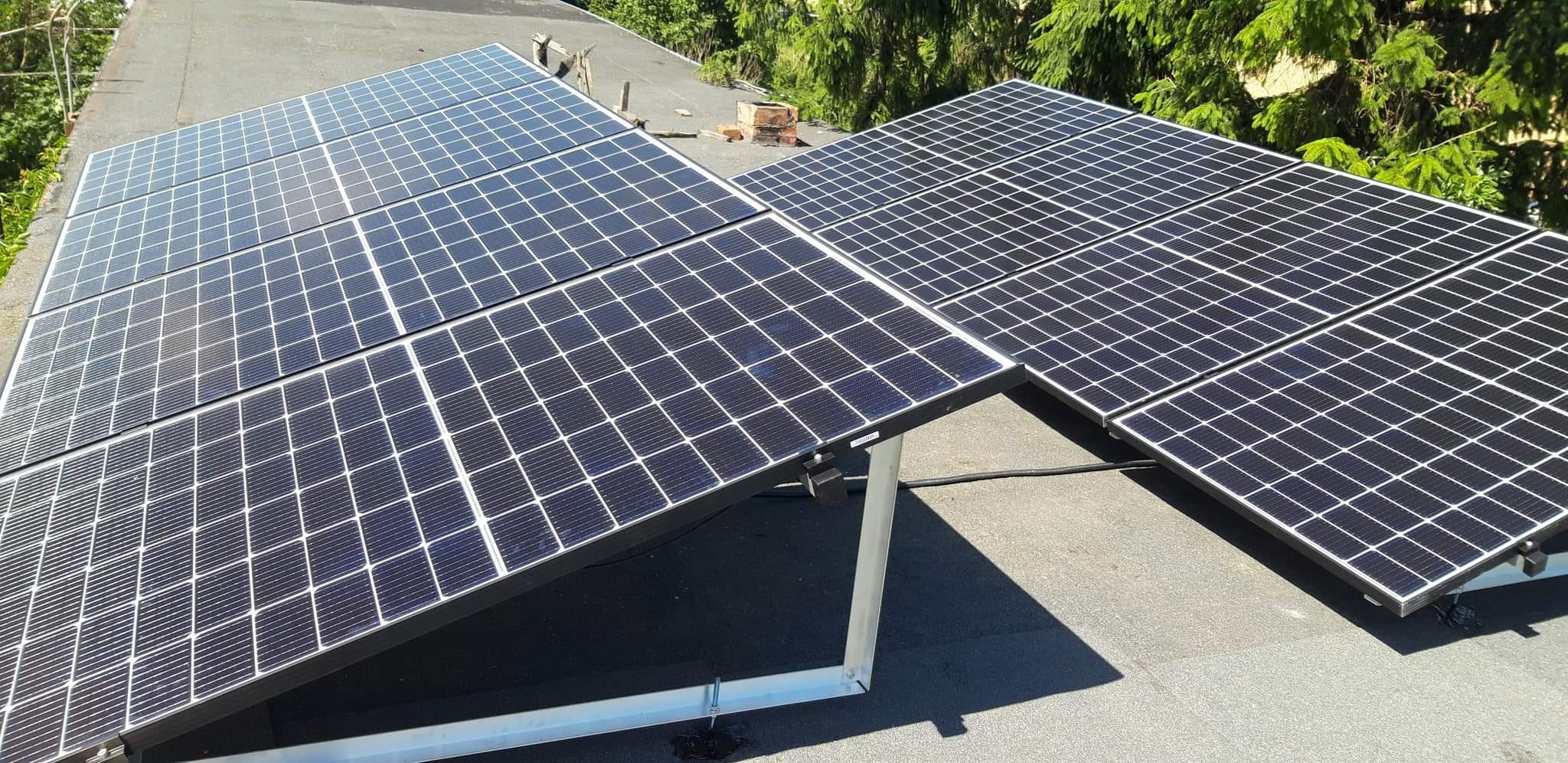 Panele fotowoltaiczne zamontowane na płaskim dachu, widoczne elementy mocujące i okablowanie, w tle zieleń drzew.