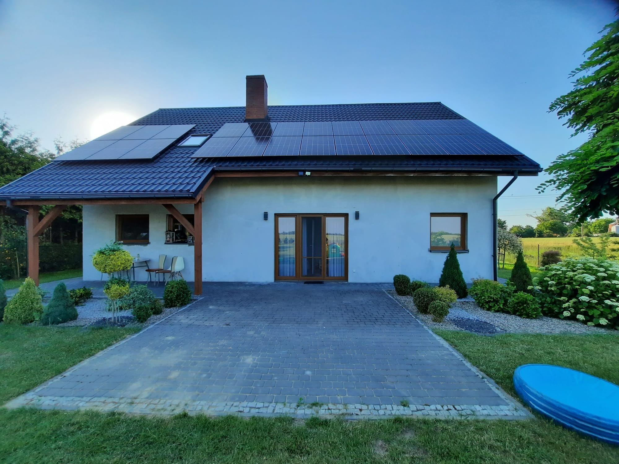 Dom jednorodzinny z panelami słonecznymi na dachu z dachówki ceramicznej, widok z poziomu chodnika z kostki brukowej, ogród z ozdobnymi krzewami i małym basenem.