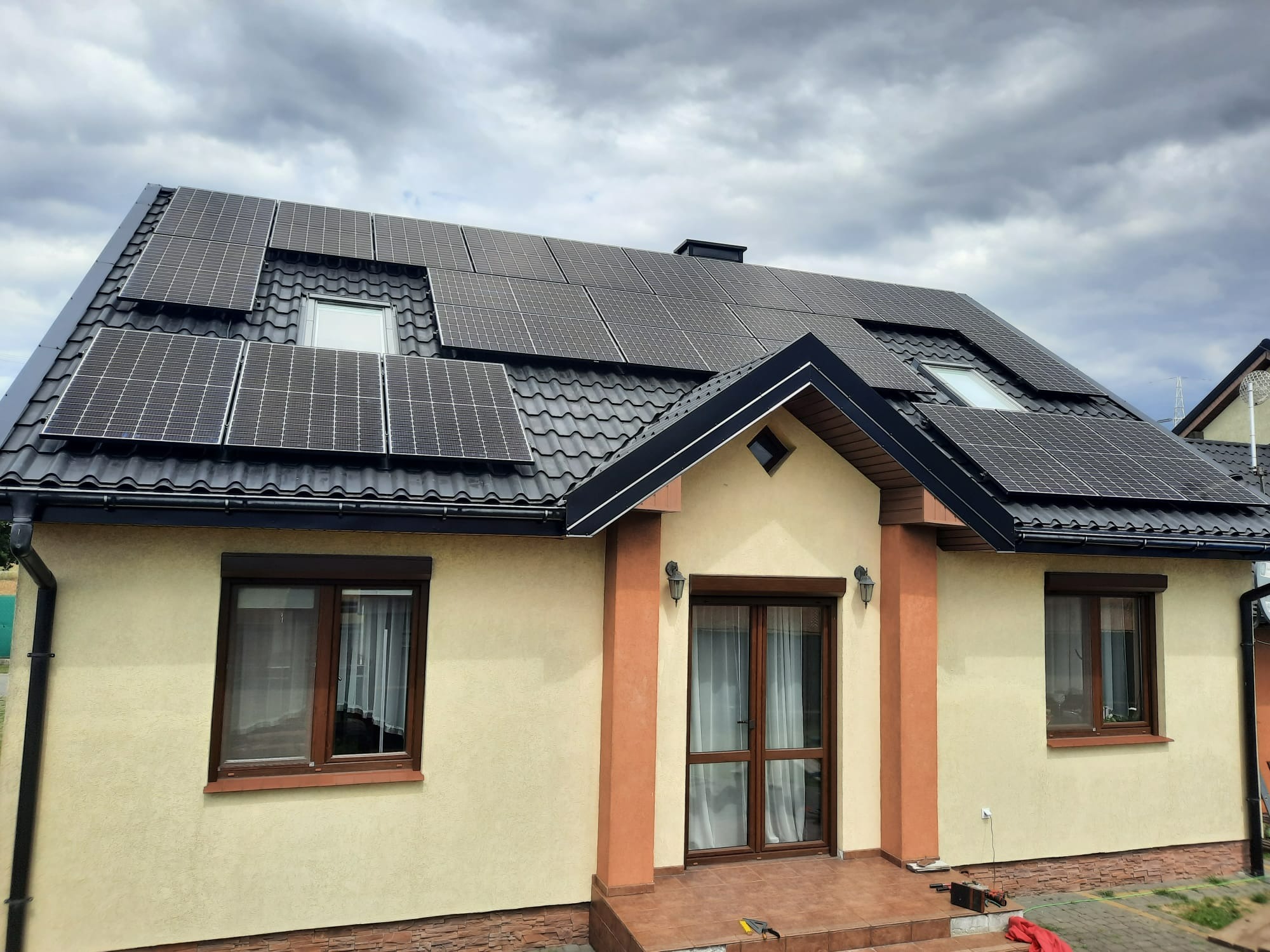 Dom jednorodzinny z panelami słonecznymi na czarnym, metalowym dachu, widoczne okno dachowe i pochmurne niebo.