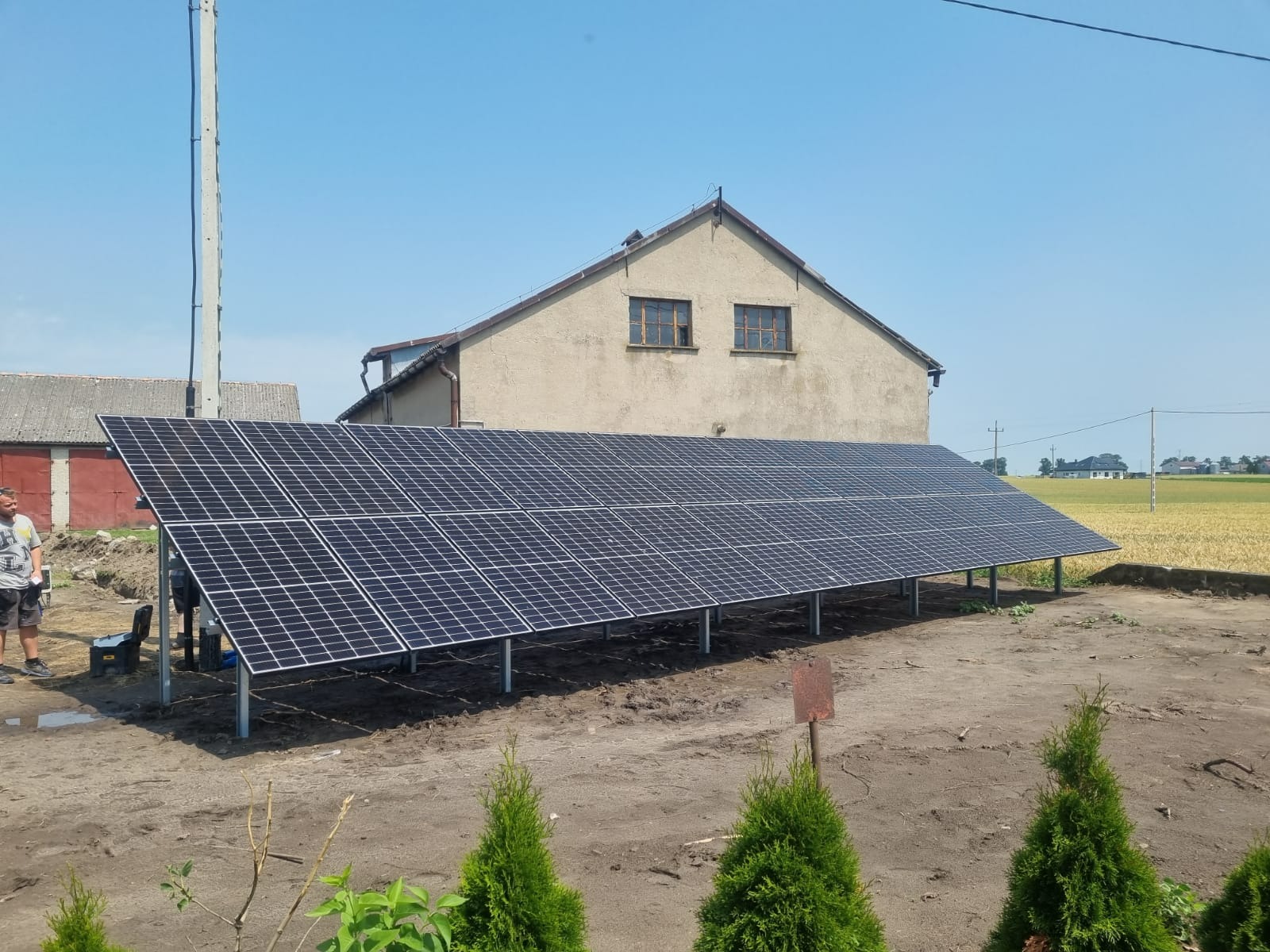 Instalacja paneli słonecznych na metalowej konstrukcji przed budynkiem gospodarczym z widocznym polem w tle.