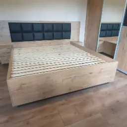 Łóżko drewniane z tapicerowanym wezgłowiem i szafkami nocnymi, widoczne w odbiciu lustrzanych drzwi szafy przesuwnej, na podłodze panele imitujące drewno.