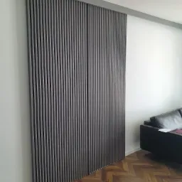 Panel ścienny z pionowych listew w kolorze ciemnego drewna, zamontowany na białej ścianie, widoczny fragment skórzanej sofy i parkiet w jodełkę.