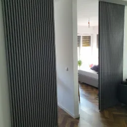 Przesuwne drzwi lamelowe w kolorze grafitowym, oddzielające dwa pomieszczenia, widok z perspektywy pokoju z parkietem w jodełkę.