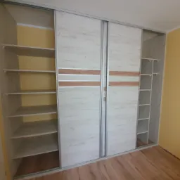 Szafa wnękowa z przesuwnymi drzwiami w odcieniach bieli i drewna, częściowo otwarta, ukazująca puste półki, na tle żółtej ściany i drewnianej podłogi.