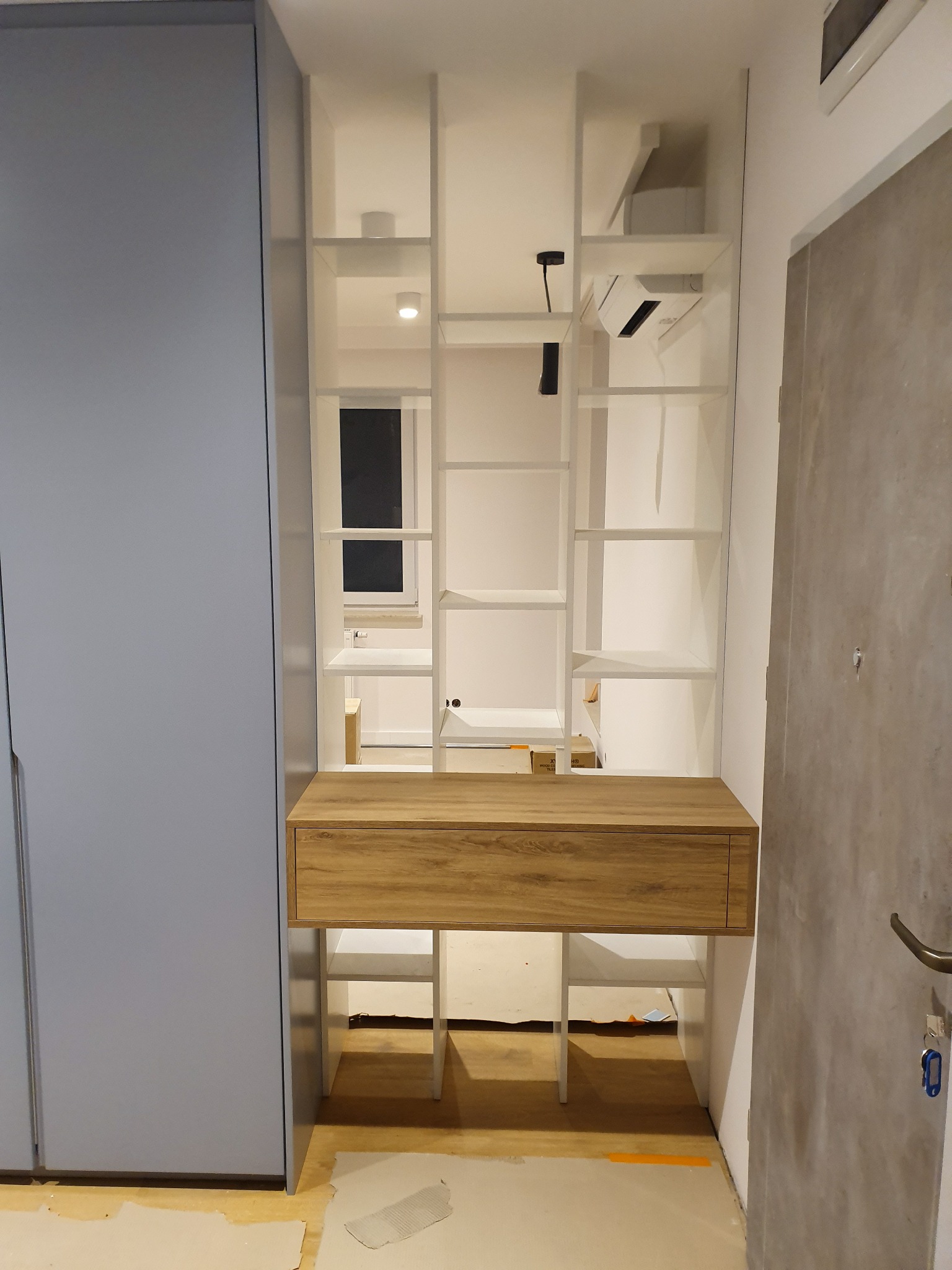 Wnęka z drewnianym blatem i szufladą, wkomponowana w biały regał z półkami, obok szafa w kolorze gołębim i szara ściana z drzwiami.