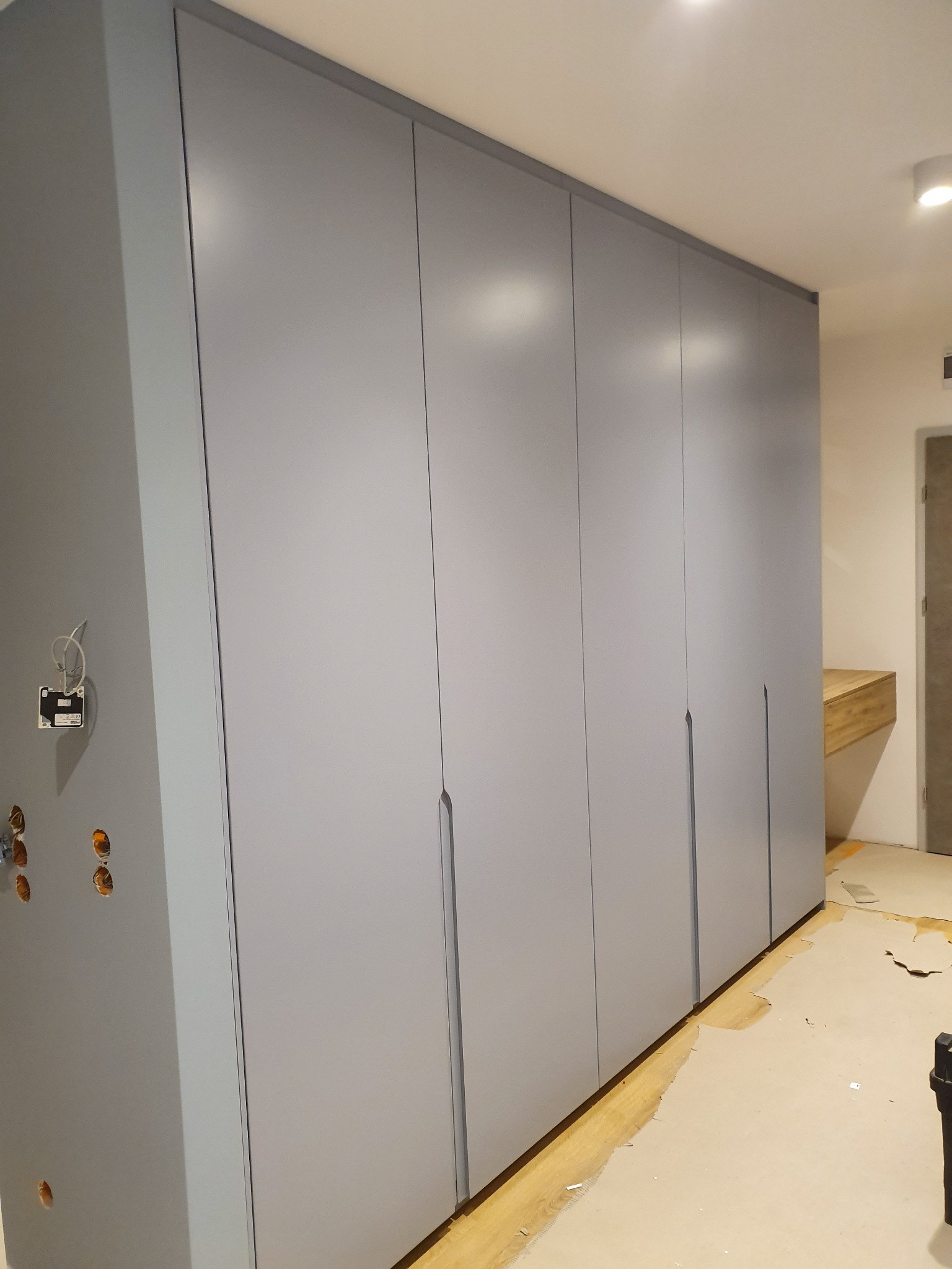 Szara, zabudowana szafa wnękowa z minimalistycznymi uchwytami, widoczne niedokończone prace wykończeniowe, w tle drewniany blat biurka.