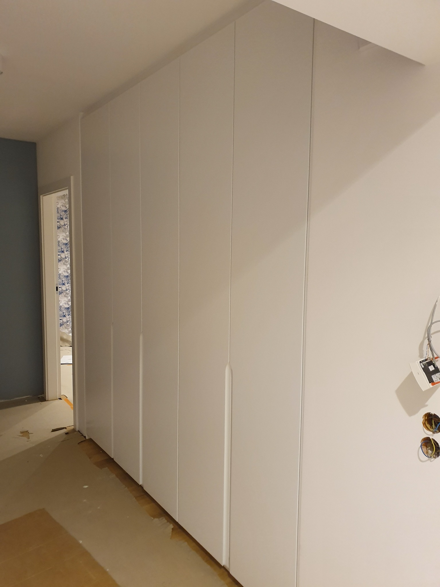 Biała szafa wnękowa z długimi, pionowymi uchwytami, widoczna na tle ściany z wystającymi kablami elektrycznymi w trakcie remontu mieszkania.