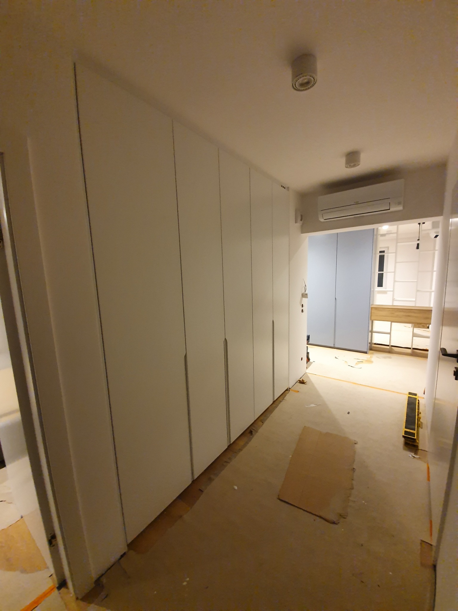 Długa, biała szafa wnękowa z minimalistycznymi uchwytami, widoczna w korytarzu podczas montażu, z zabezpieczonymi podłogami i widoczną przestrzenią garderoby w tle.