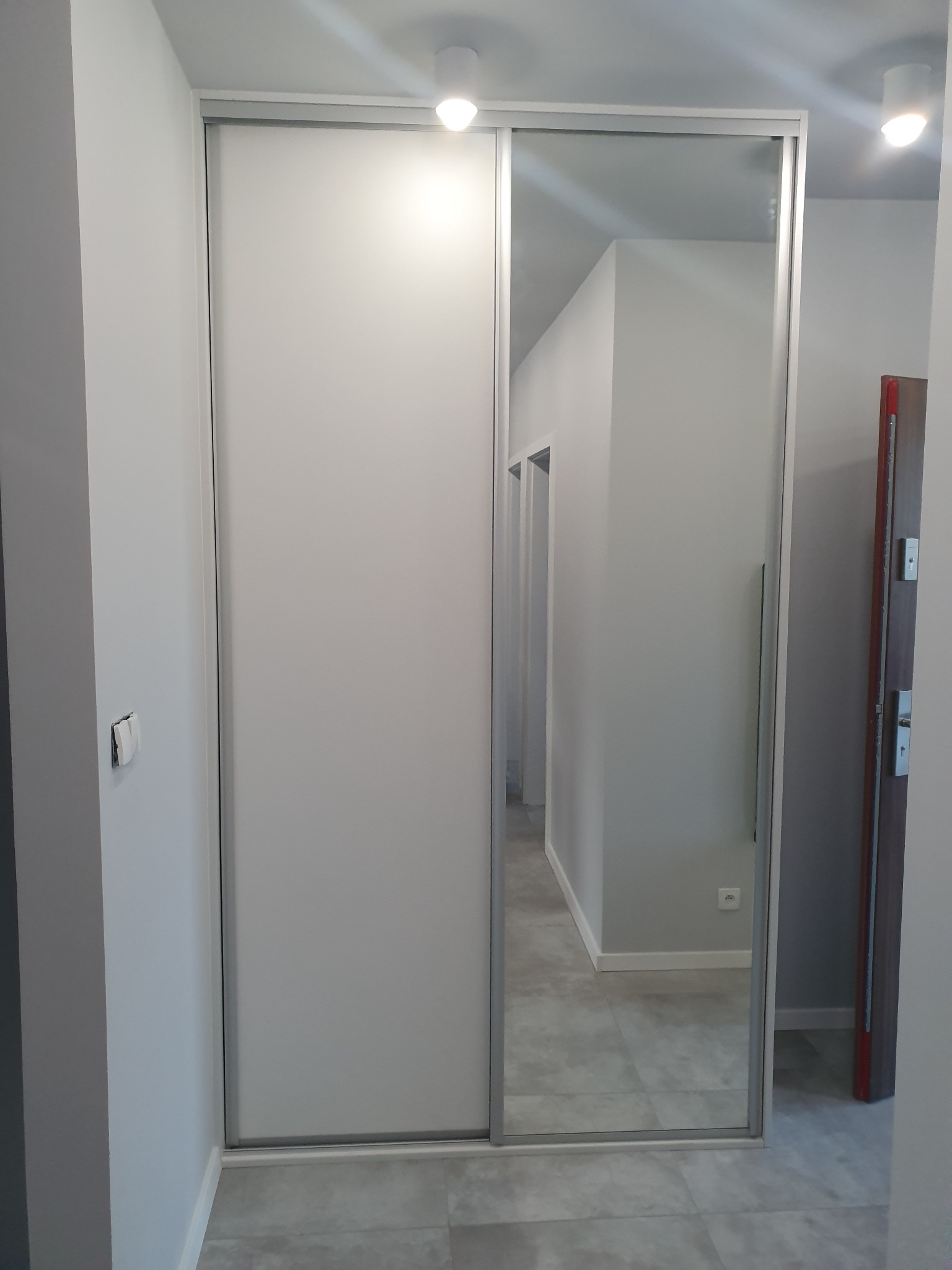 Nowoczesna szafa wnękowa z przesuwnymi drzwiami, jedna część z białym panelem, druga z lustrem odbijającym wnętrze mieszkania. Widoczna szara podłoga i białe ściany.