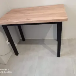 Stół z blatem imitującym jasne drewno i czarnymi, metalowymi nogami w minimalistycznym stylu, ustawiony na szarej podłodze.