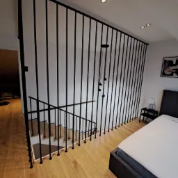 Nowoczesna, czarna balustrada schodowa z pionowych prętów, oddzielająca schody od sypialni z łóżkiem i lampką nocną.