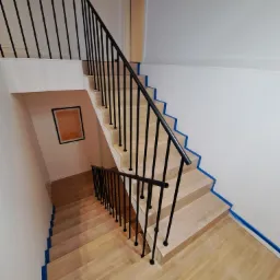 Wysoki kąt widzenia na drewniane schody z czarną, minimalistyczną balustradą; ściany pomalowane na biało z niebieską taśmą malarską wzdłuż krawędzi schodów i ścian.