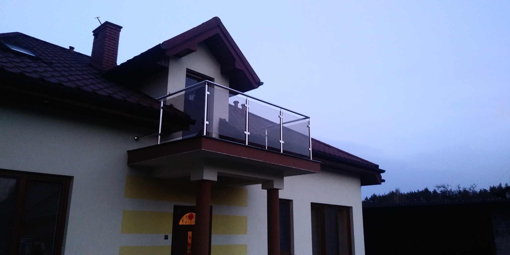 Nowoczesny balkon z szklaną balustradą na piętrze domu jednorodzinnego z brązowym dachem i kominem.