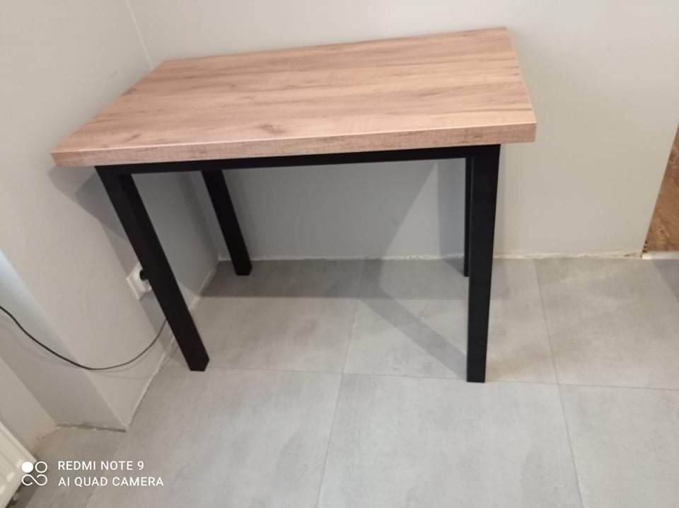 Stół z blatem imitującym jasne drewno i czarnymi, metalowymi nogami w minimalistycznym stylu, ustawiony na szarej podłodze.