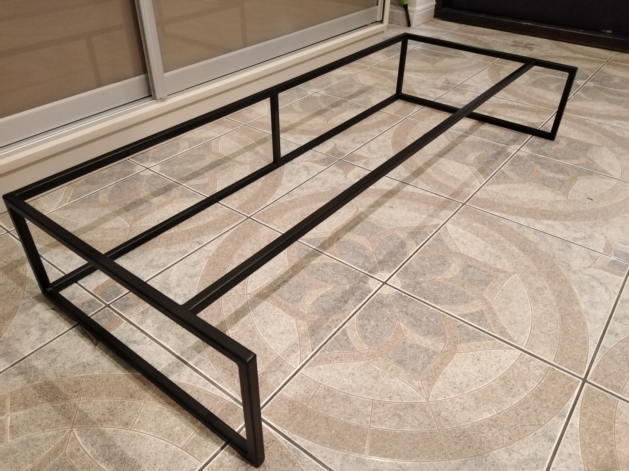 Czarna, prostokątna rama ze spawanych profili stalowych, leżąca na podłodze z płytek ceramicznych z wzorem rozetowym. Widoczne miejsce spawania profili.