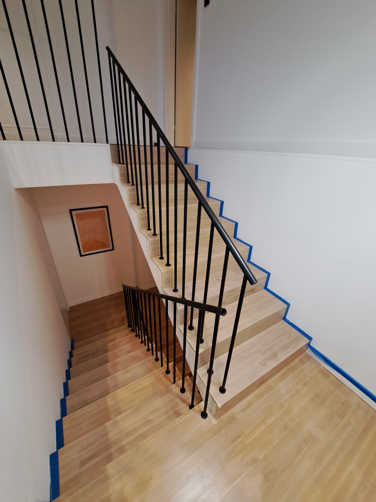 Wysoki kąt widzenia na drewniane schody z czarną, minimalistyczną balustradą; ściany pomalowane na biało z niebieską taśmą malarską wzdłuż krawędzi schodów i ścian.