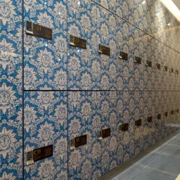 Rząd szafek z niebieskim, kwiecistym wzorem, zabezpieczonych zamkami szyfrowymi, w nowoczesnym wnętrzu z drewnianymi panelami i szarą podłogą.