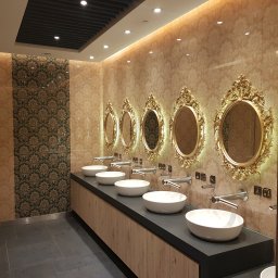 A.M.G. KATOWICE SPÓŁKA Z OGRANICZONĄ ODPOWIEDZIALNOŚCIĄ - Luksusowa łazienka publiczna z rzędem owalnych umywalek i ozdobnych luster w złotych ramach na tle wzorzystej tapety w ciepłych odcieniach.