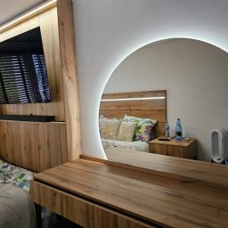 A.M.G. KATOWICE SPÓŁKA Z OGRANICZONĄ ODPOWIEDZIALNOŚCIĄ - Nowoczesna sypialnia z toaletką i okrągłym lustrem z podświetleniem LED, odbijającym łóżko z drewnianym zagłówkiem i szafki nocne. Drewniane meble w ciepłych odcieniach.