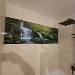 A.M.G. KATOWICE SPÓŁKA Z OGRANICZONĄ ODPOWIEDZIALNOŚCIĄ - Panel szklany z motywem wodospadu w łazience, nadający naturalny akcent. Beżowe płytki i czarna armatura prysznicowa. Nowoczesne wykończenie.