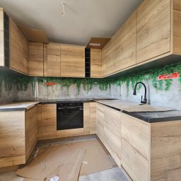 A.M.G. KATOWICE SPÓŁKA Z OGRANICZONĄ ODPOWIEDZIALNOŚCIĄ - Jasna, drewnopodobna zabudowa kuchenna w kształcie litery U, z czarnym blatem i motywem roślinnym na ścianie. Widoczne kartony zabezpieczające podłogę.