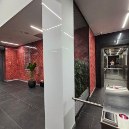 A.M.G. KATOWICE SPÓŁKA Z OGRANICZONĄ ODPOWIEDZIALNOŚCIĄ - Nowoczesne wnętrze biurowca z przeszkloną kolumną i elementami dekoracyjnymi. Widoczny fragment windy i roślinność w tle. Oświetlenie LED podkreśla detale.