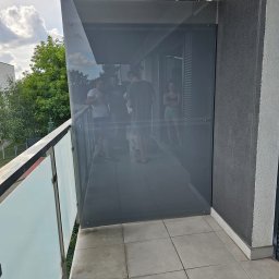 A.M.G. KATOWICE SPÓŁKA Z OGRANICZONĄ ODPOWIEDZIALNOŚCIĄ - Szklana zabudowa balkonu z widocznymi mocowaniami, odbijająca sylwetki osób wewnątrz. Nowoczesne rozwiązanie architektoniczne z szarymi płytkami podłogowymi.