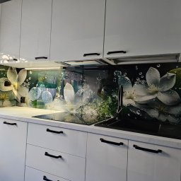 A.M.G. KATOWICE SPÓŁKA Z OGRANICZONĄ ODPOWIEDZIALNOŚCIĄ - Biała kuchnia z błyszczącymi szafkami i panelem szklanym z motywem kwiatowym nad blatem. Czarne uchwyty i armatura. Widoczne odbicie osoby w szkle.