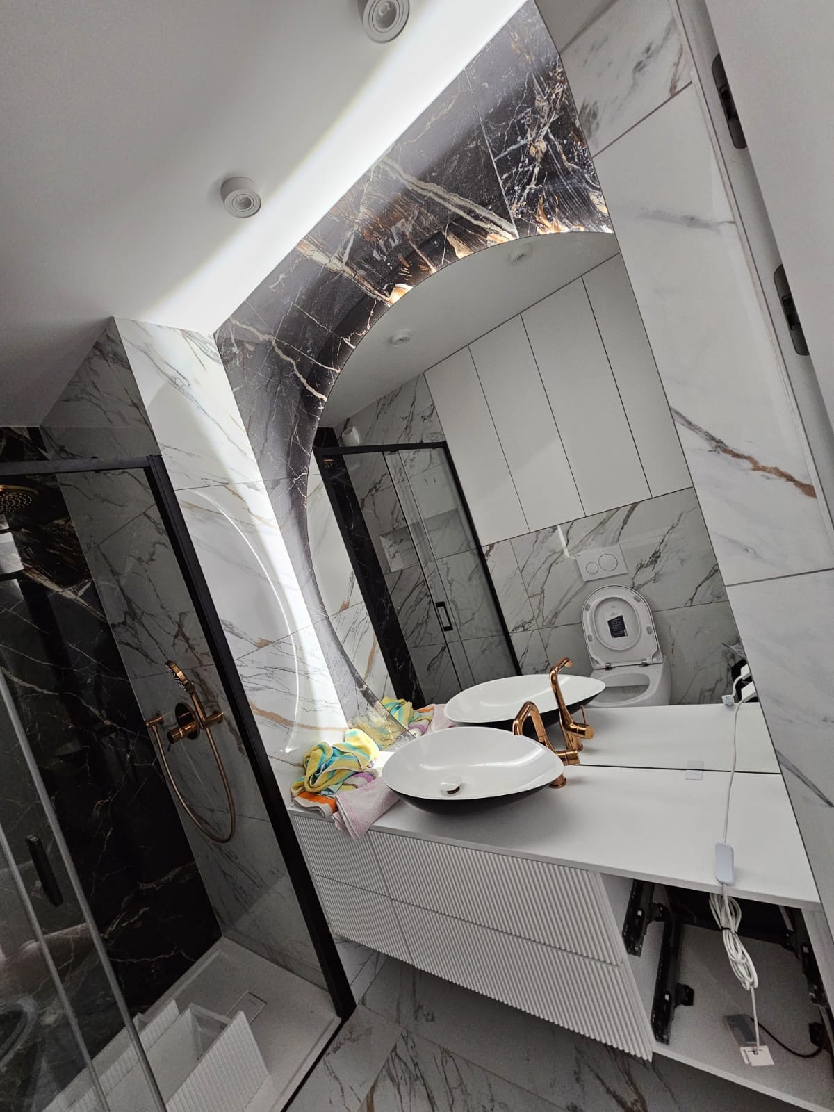 Nowoczesna łazienka z marmurowymi płytkami, kabiną prysznicową typu walk-in, podwójną umywalką i dużym, owalnym lustrem z podświetleniem.