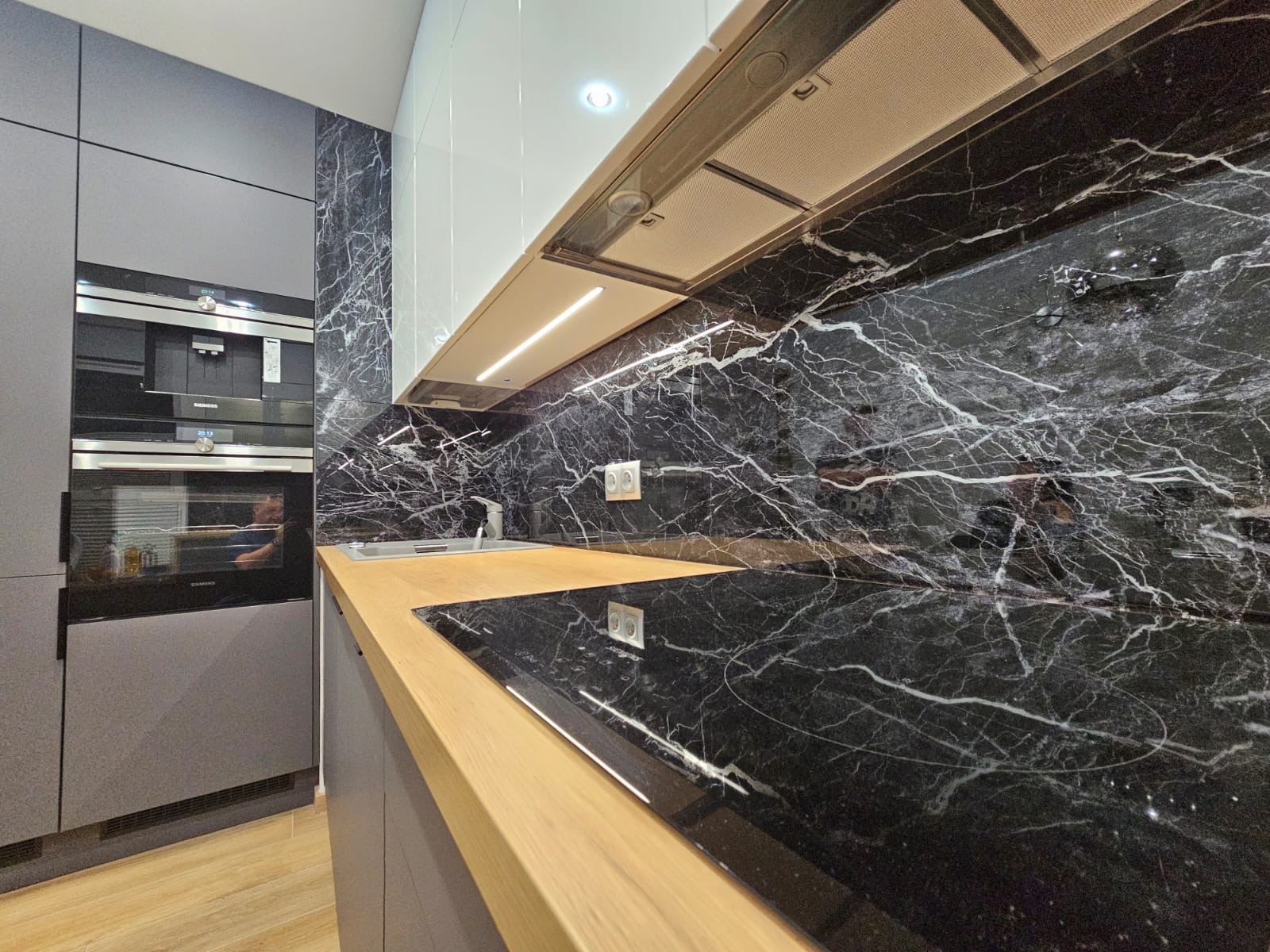 Nowoczesna kuchnia z blatem z jasnego drewna i czarnym marmurem na ścianie. Widoczny zlew, płyta indukcyjna i zabudowane sprzęty AGD.