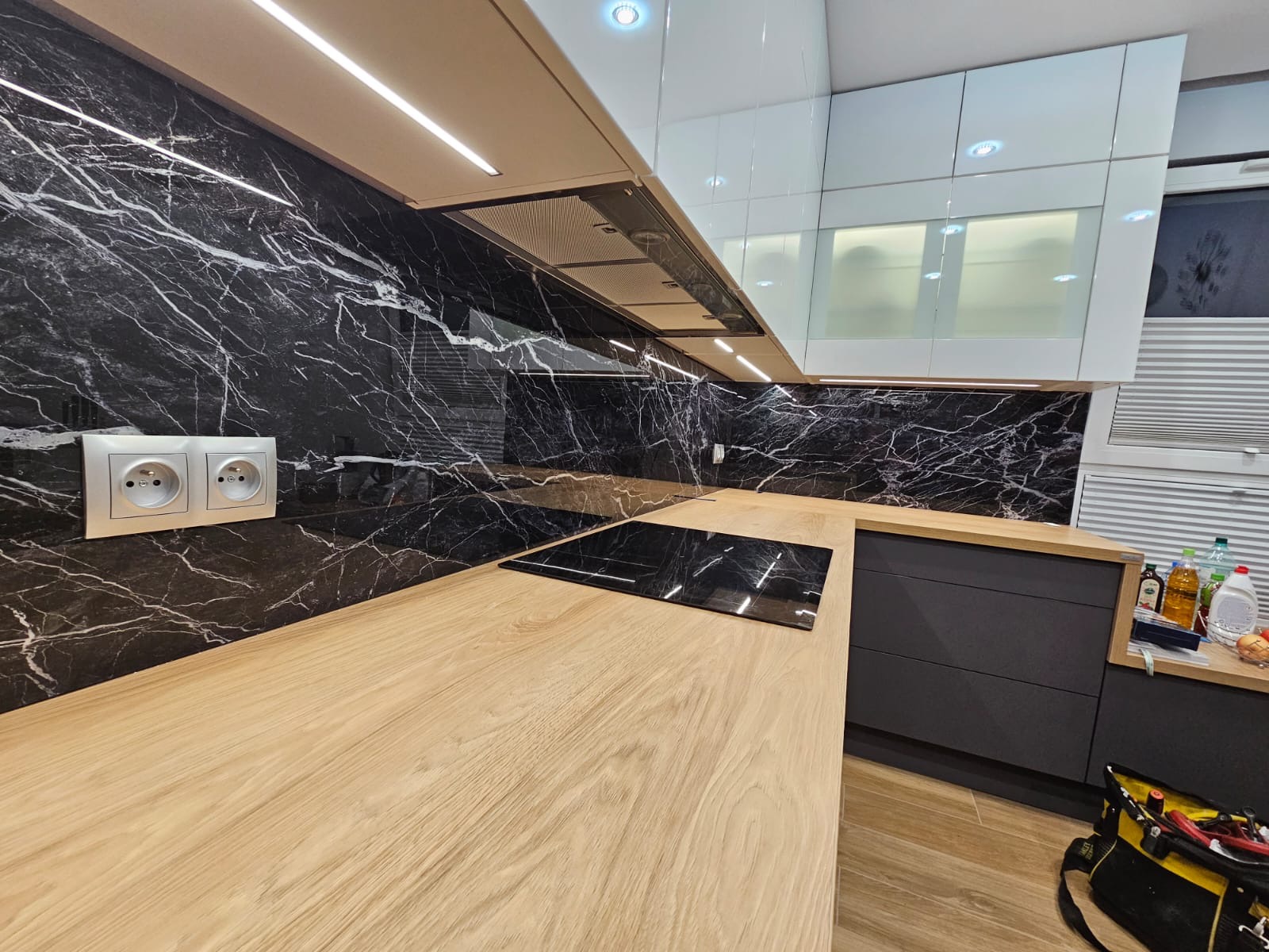 Nowoczesna kuchnia z blatem z jasnego drewna i czarnym marmurem na ścianie. Widoczne gniazdka elektryczne i płyta indukcyjna. Oświetlenie LED pod szafkami.
