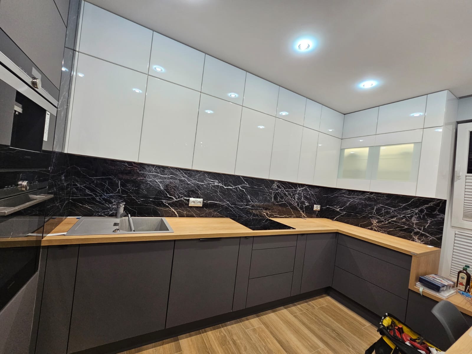 Nowoczesna kuchnia z szarymi szafkami dolnymi, białymi górnymi i blatem z drewnopodobnego laminatu. Czarny panel ścienny z białym marmurkowym wzorem.