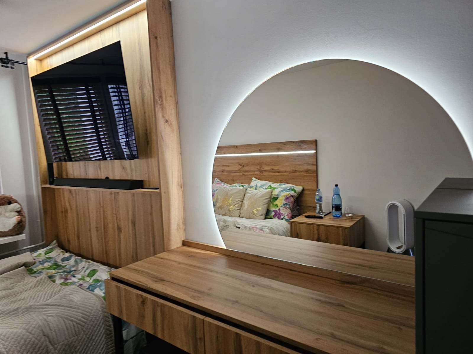 Nowoczesna sypialnia z toaletką i okrągłym lustrem z podświetleniem LED, odbijającym łóżko z drewnianym zagłówkiem i szafki nocne. Drewniane meble w ciepłych odcieniach.