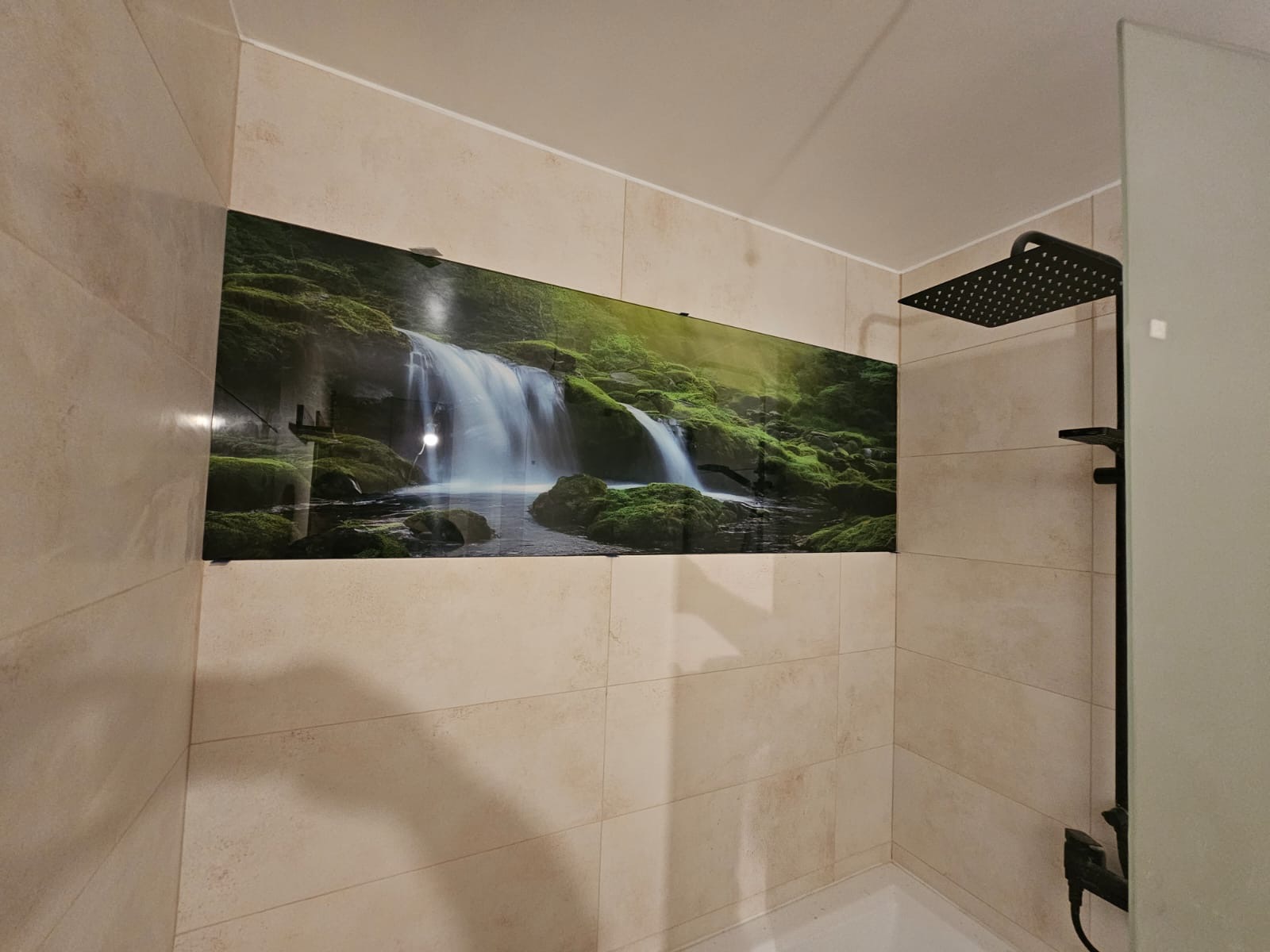 Panel szklany z motywem wodospadu w łazience, nadający naturalny akcent. Beżowe płytki i czarna armatura prysznicowa. Nowoczesne wykończenie.