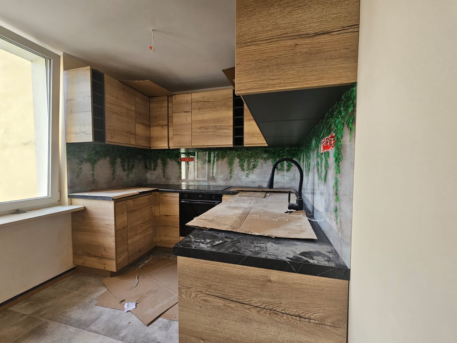 Kuchnia z drewnianymi szafkami i czarnym blatem, ściana z motywem roślinnym. Widoczne kartony na podłodze, sugerujące prace wykończeniowe. Ujęcie z perspektywy wnętrza.