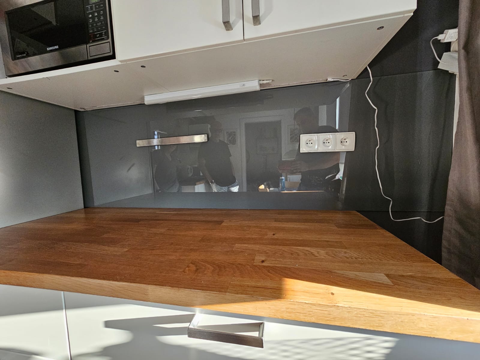 Szklany panel nad blatem kuchennym, z widocznymi gniazdkami elektrycznymi i odbiciem osób w tle. Blat drewniany, oświetlenie podszafkowe.