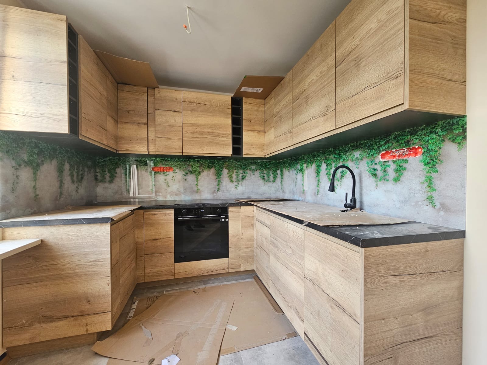 Jasna, drewnopodobna zabudowa kuchenna w kształcie litery U, z czarnym blatem i motywem roślinnym na ścianie. Widoczne kartony zabezpieczające podłogę.