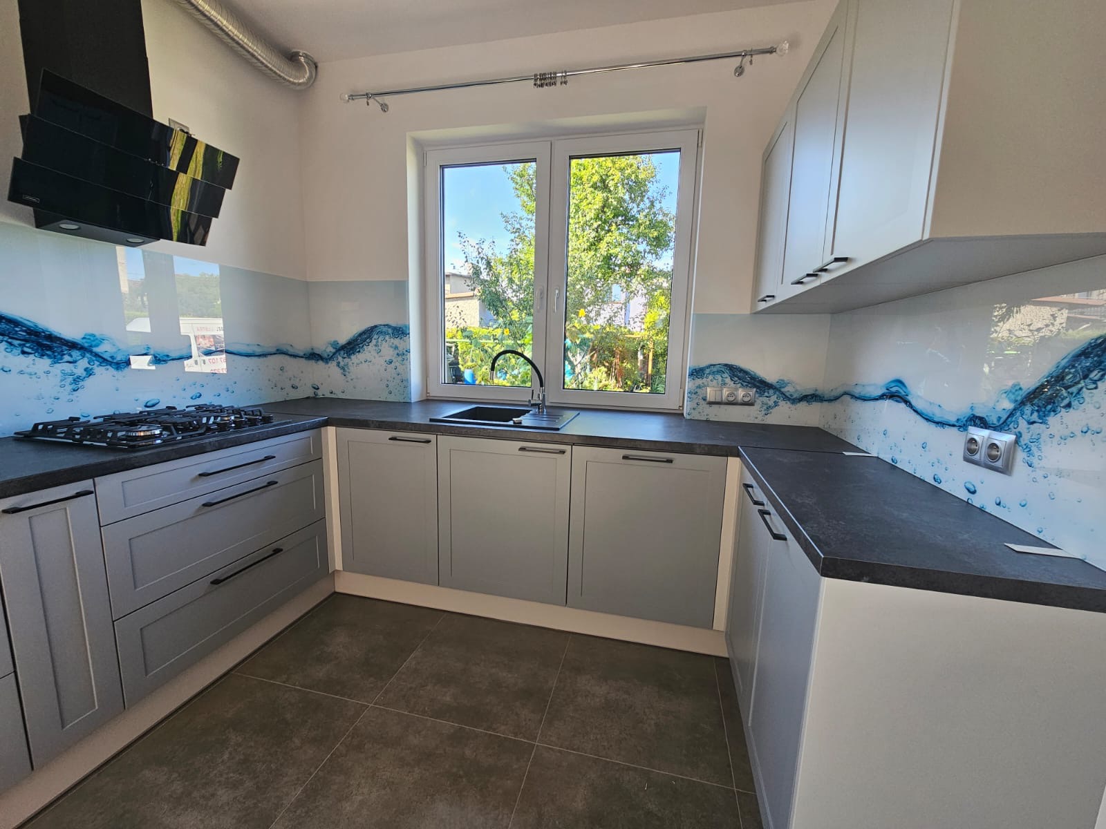 Jasna kuchnia z szarymi szafkami i blatem z ciemnego kamienia. Nad blatem panel szklany z motywem wody. Widok na zewnątrz przez okno.