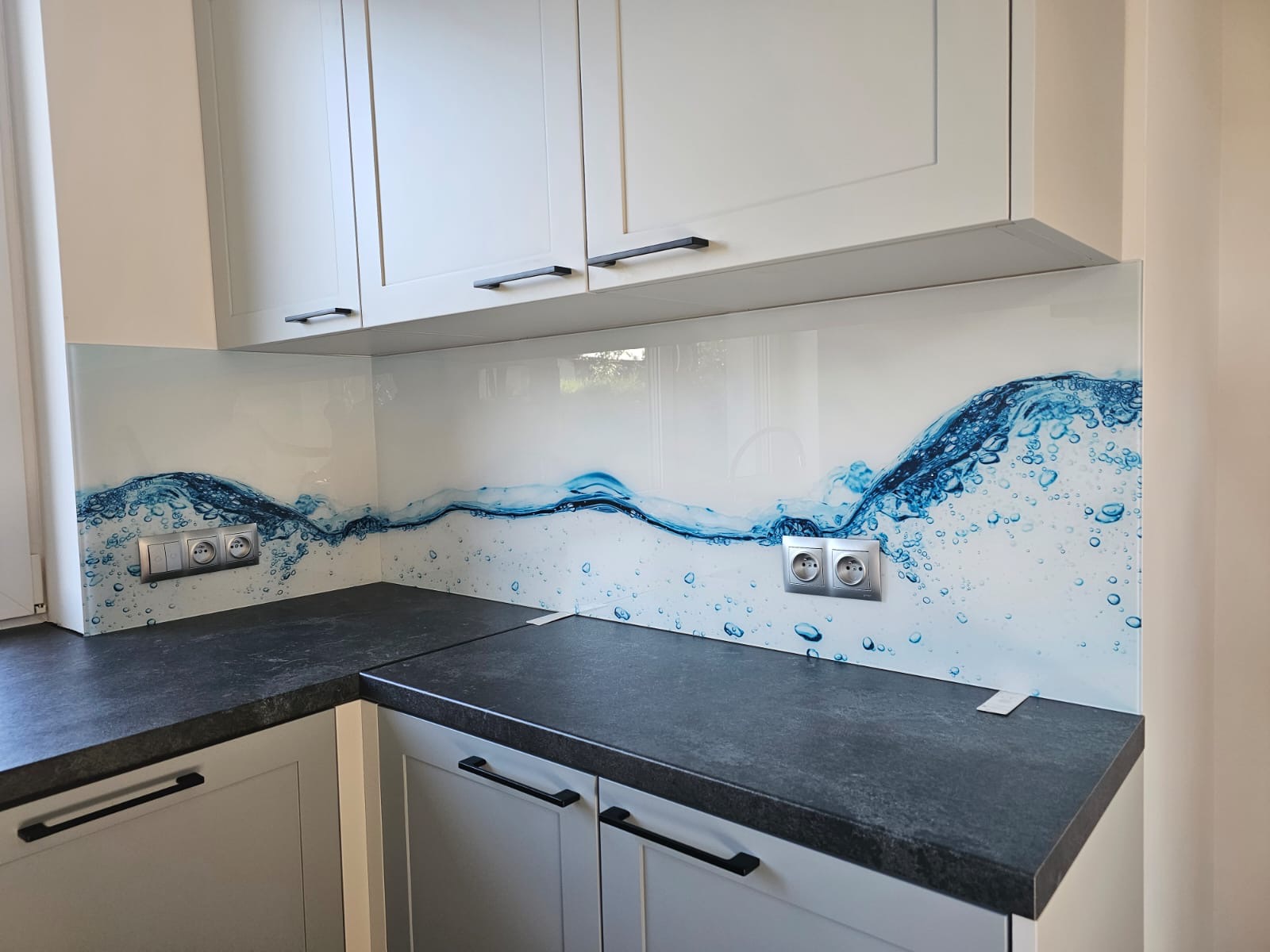 Szklany panel kuchenny z motywem wody, zamontowany między blatem a szafkami. Widoczne gniazdka elektryczne i fragment blatu z szafkami w dolnej części kadru.
